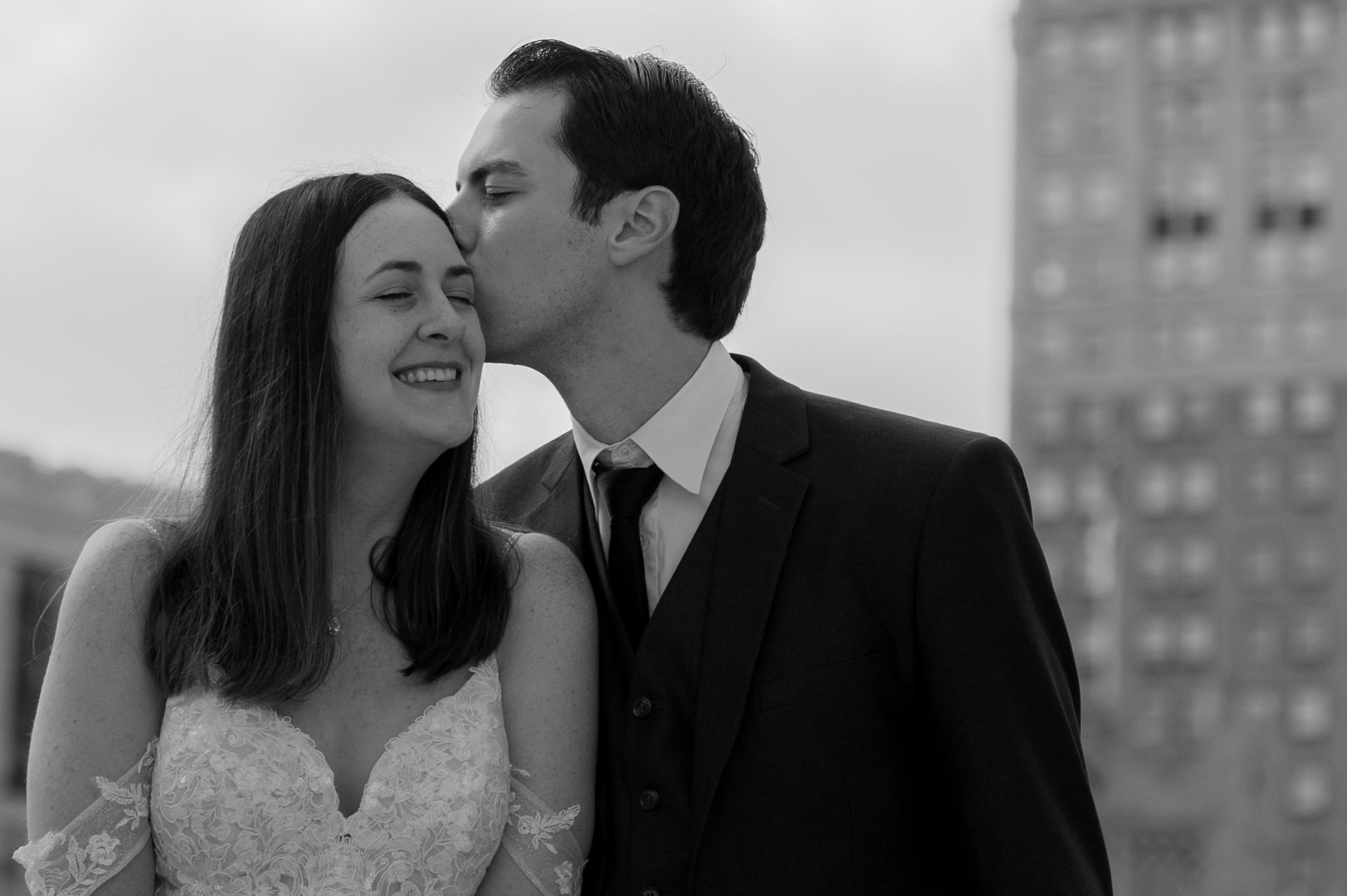 Black and white photo of groom kissing the bride's temple, in an urban setting